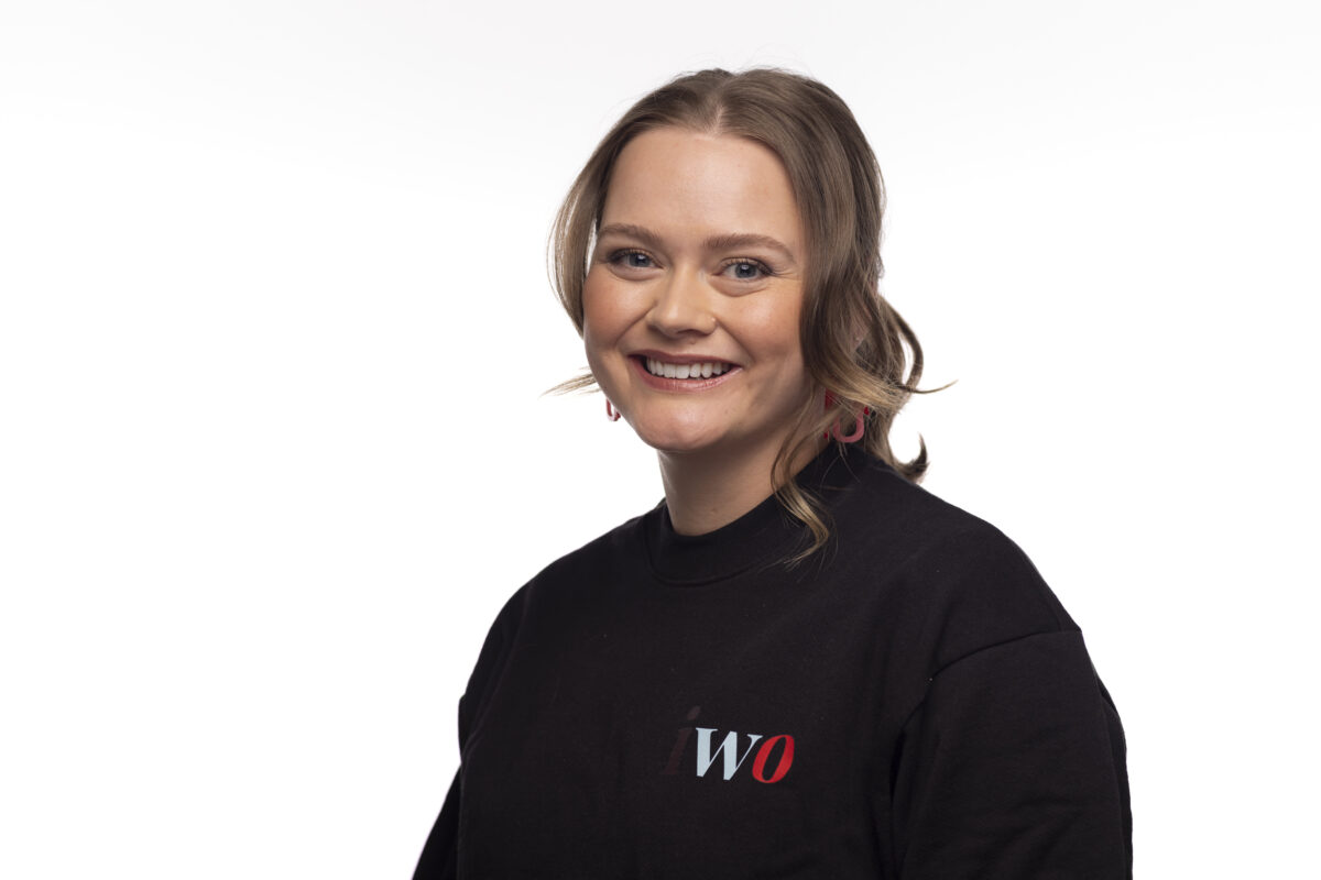 Aiyana- Indigenous Women Outdoors' Executive Director. Sitting and smiling with her hair in a ponytail, in a black long sleeve with the IWO logo on its front.